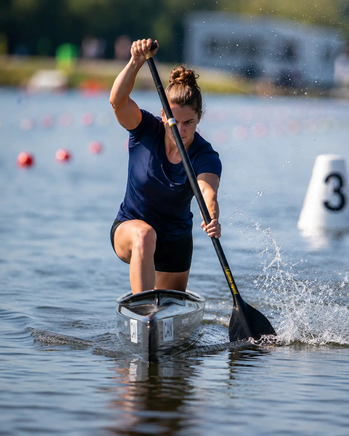 Athletes competing in a canoe sprint event, photographed from the water's edge on a flat-water race course