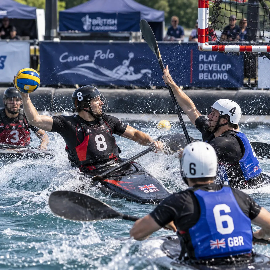 Canoe polo players in a goalmouth scramble, reaching for the ball beside a red-and-white striped goal in full-face helmets and GBR vests