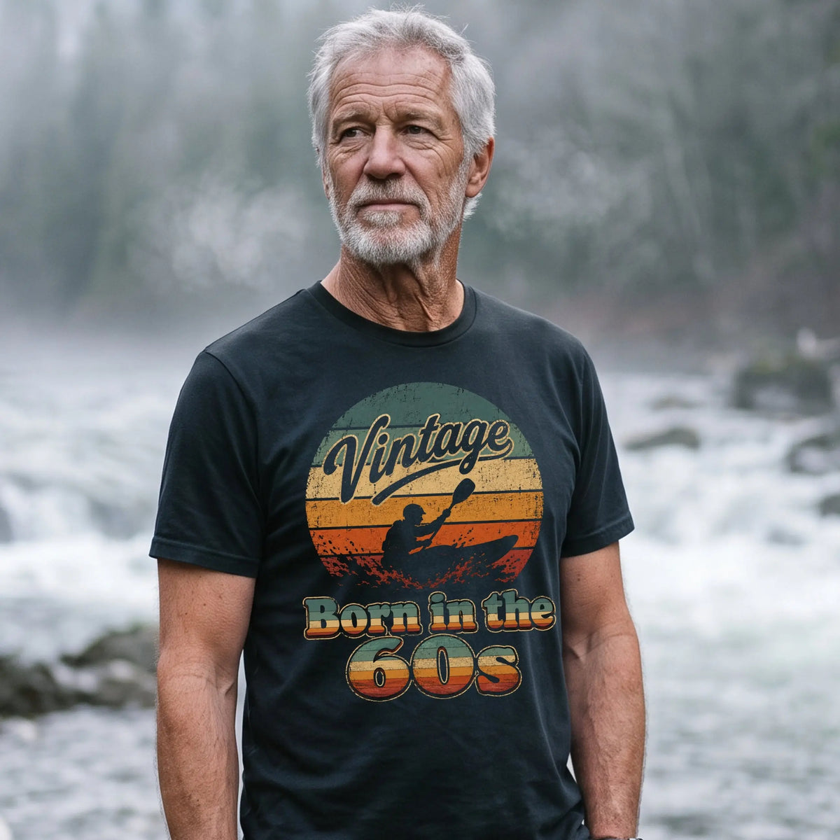 An older man with gray hair and a beard stands on a rocky riverside in a misty forest setting, wearing a black graphic t-shirt with a retro sunset design that reads 'Vintage Born in the GOS'. A green kayak and paddle are visible on the shore next to him.