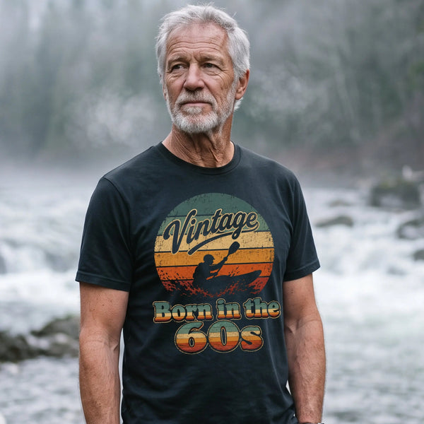 An older man with gray hair and a beard stands on a rocky riverside in a misty forest setting, wearing a black graphic t-shirt with a retro sunset design that reads 'Vintage Born in the GOS'. A green kayak and paddle are visible on the shore next to him.