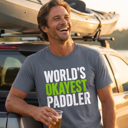 Man laughing in a grey World's Okayest Paddler t-shirt, holding a drink beside his car with a kayak on the roof rack after a paddle — Vector Paddle humour tee