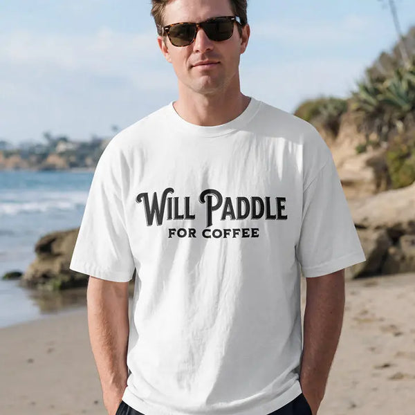 A man standing on a sandy beach wearing a white crewneck t-shirt with the black text 'Will PADDLE FOR COFFEE' and black shorts.