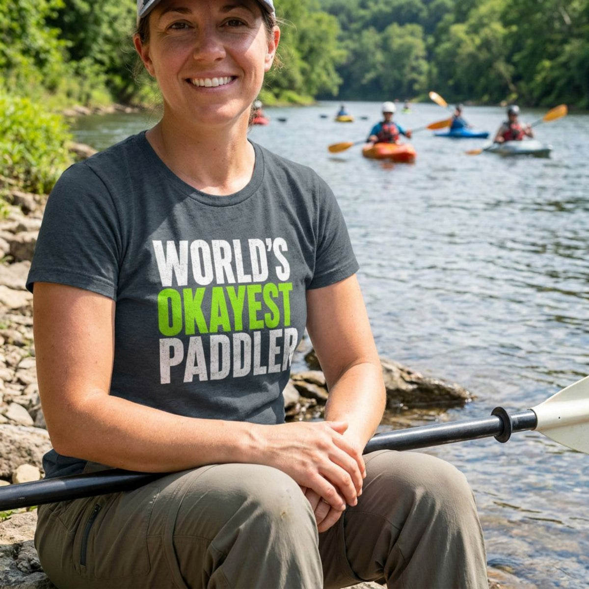 Woman smiling streamside in a grey World's Okayest Paddler fitted tee, paddle across her knees, kayakers passing on the river behind her — Vector Paddle women's paddling humour tee