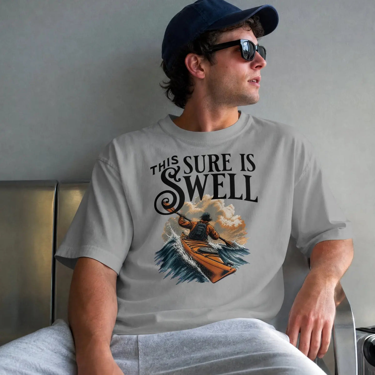 A man wearing a light gray graphic t-shirt with the text 'THIS SURE IS WELL' and a surfer illustration, sitting on a metal bench in an airport lounge.