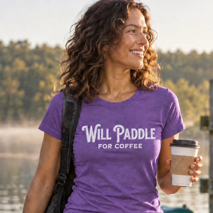 A woman walking on a wooden dock by a lake wearing a purple Vector Paddle t-shirt that reads 'Will Paddle For Coffee' and holding a coffee cup. Kayaks and a paddle are visible in the background, with the Vector Paddle logo in the top right corner.